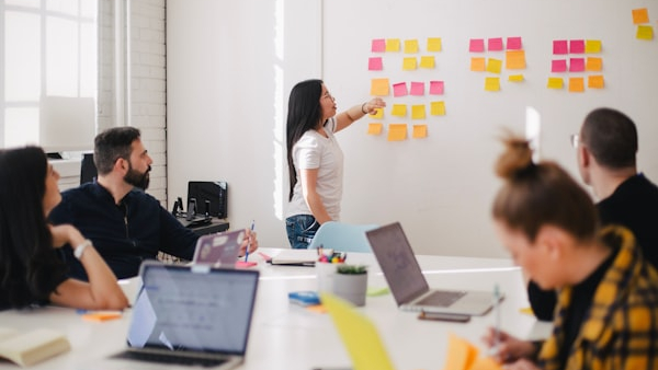 Team in a bright meeting room collaborates around a table as a woman points to colorful sticky notes on the wallwall edge)